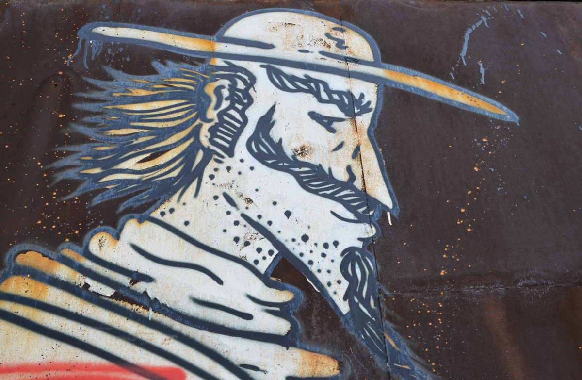 street art drawing of a caricature of a man, long pointy chin with beard, long hair in back, flat hat with large brim