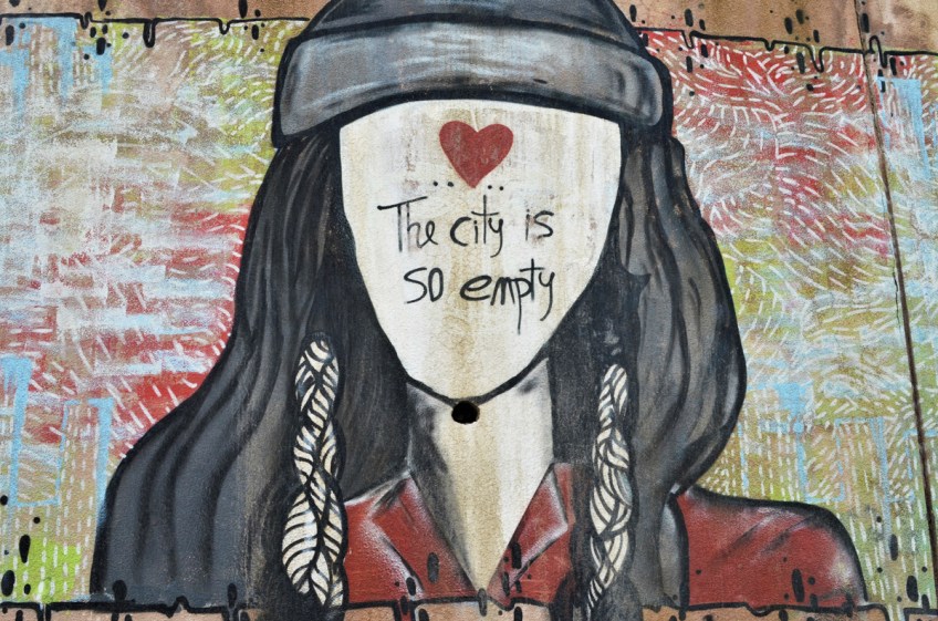 street art painting of a young woman with long black hair with a head band tied around her head, black face, no facial features, instead a red heart on her forehead and the words The city is so empty