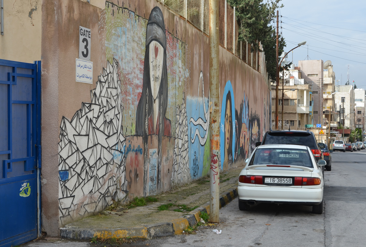 blue gate on the left, cars parked in front of, wall with street art, houses and other buildings in the background