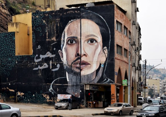 mural by herakut in Amman, very large face that is divided vertically down the middle, male on one side, female on the other, 4 storeys high, black background, man with beard and small moustache 