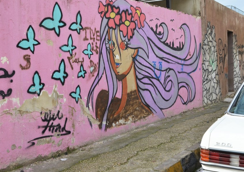mural on pink background of a young woman with long purple hair, flower crown on her head, and blue butterflies flying nearby, head and shoulders