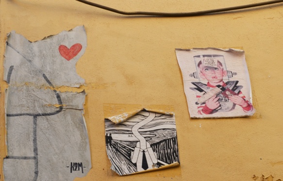 3 paper paste ups on a yellow concrete wall, all three slightly faded and peeling at the edges, one is a K2M charaacter with a red heart, another is a black and white drawing similar to Edvard Munsch's Scream painting, and the last is a drawing of a young boy with ray gun and old fashioned idea of a space uniform 
