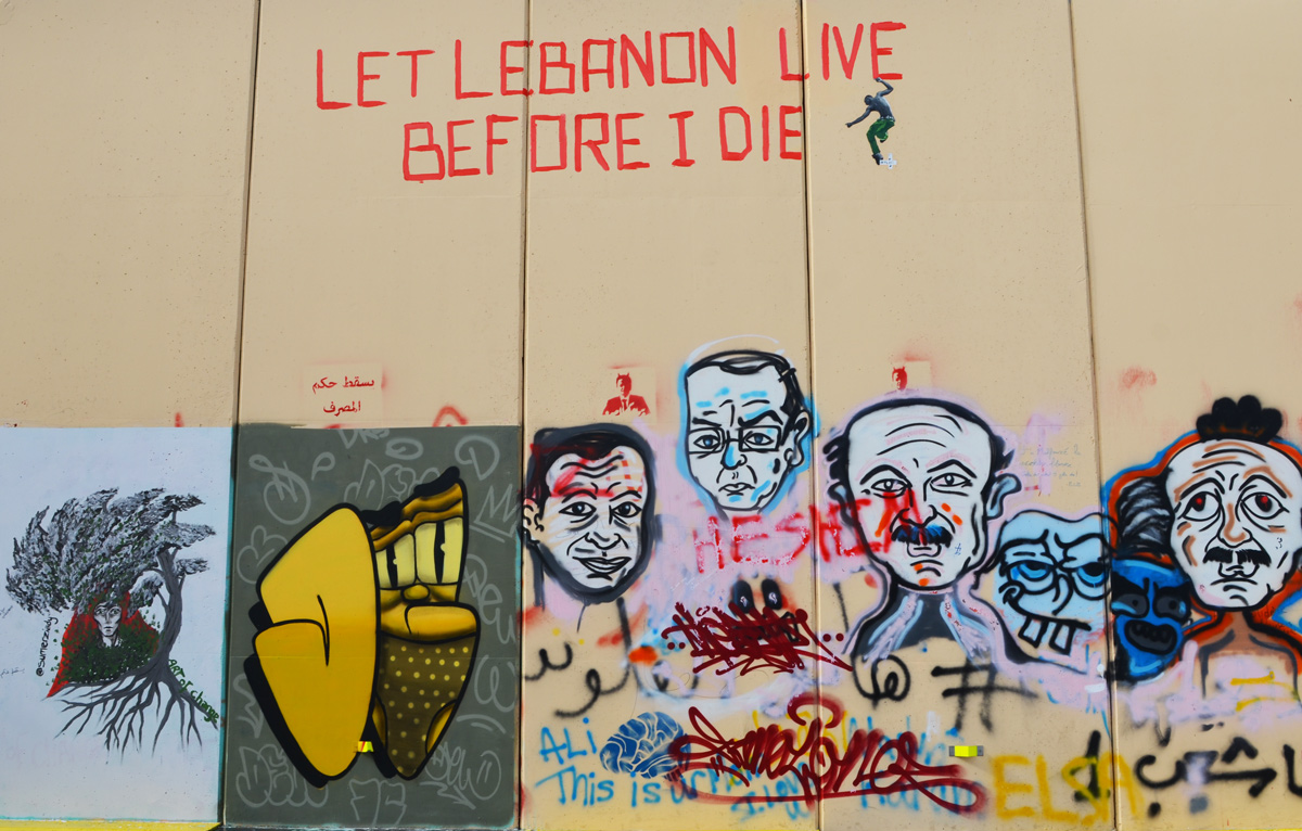 light brown temporary wall outside, made of panels, now home to a revolution wall of art and graffiti, white faces, a poster by sumerziady and the words let Lebanon live before I die 