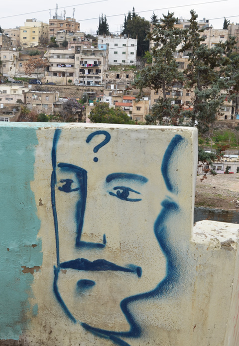 a blue line drawing of a man's face with a question mark on his forehead, painted on a short wall, view of Amman in the background