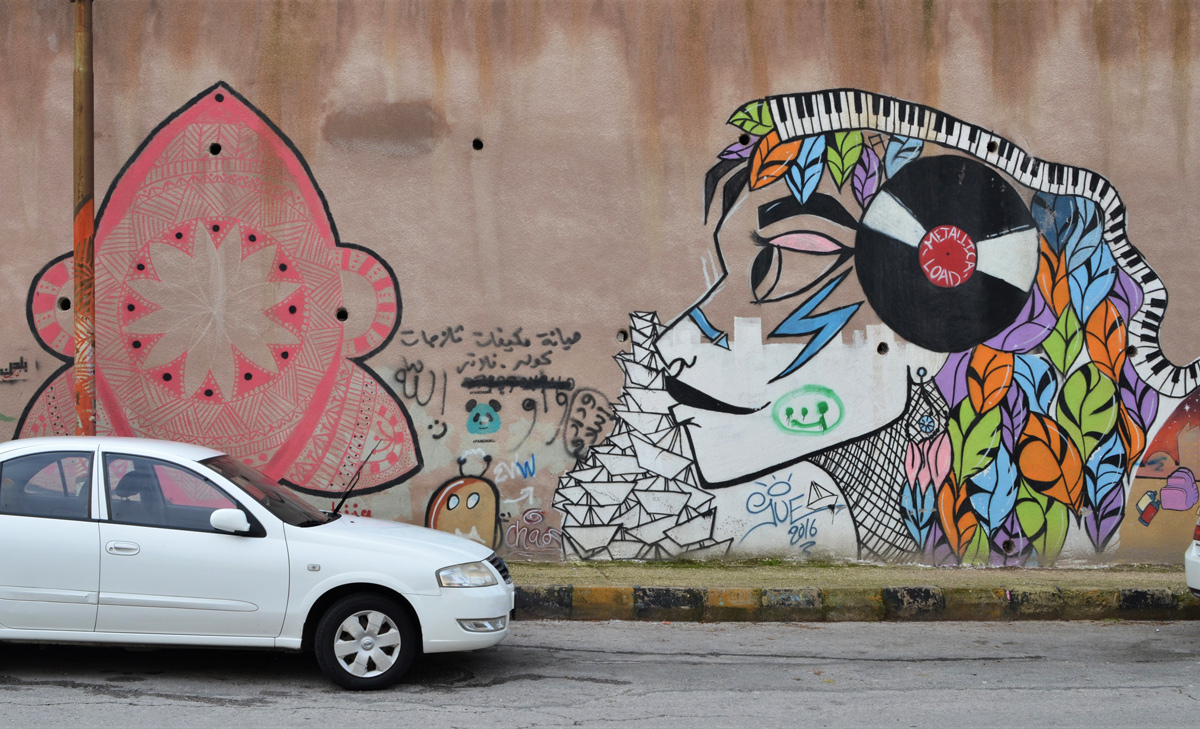 two white cars parked in front of a wall of Terra Sancta college with two murals on it, one by Suha Sultan with a woman with a Metallica Load vinyl record on the side of her head, and the other a pink shape