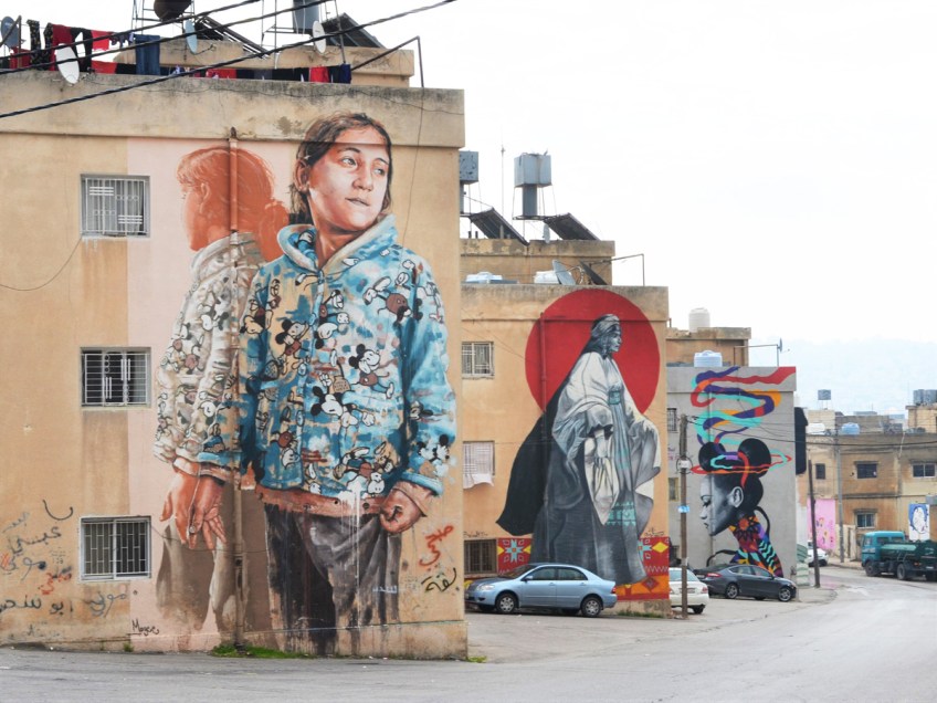 three large murals in a row, in the foreground is one of a girl by Finton Magee, the next is an old woman with a red circle in the background and the third is a colourful woman with ribbons by Dina Saadi