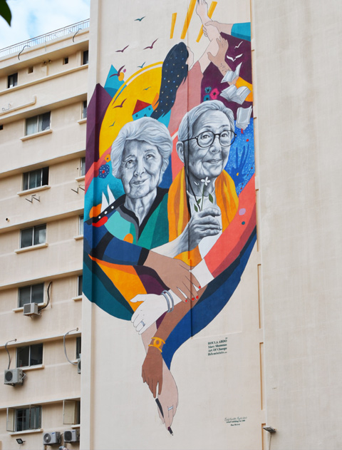 a mural on the side of a building of two older women, Emily Nasrallah and Huguette Caland, with arms linked with extra arms, coloured ribbons winding between and around them 