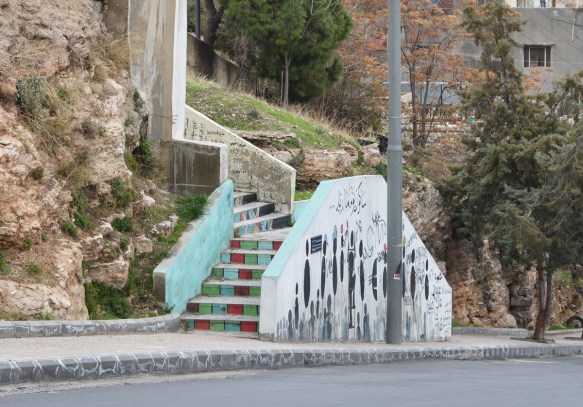 brightly coloured stairs that end with a a white wall with paintings of people made from circles and ovals with stick arms and legs, all doing something