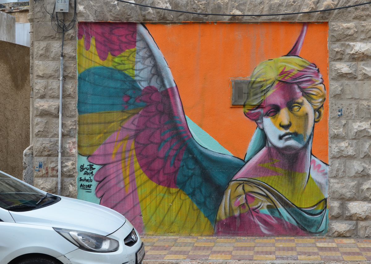 woman's head and shoulders on an orange background, mural by Suhaib Attar, woman is in blue, pink, and yellow splotches. She has wings in the same colour