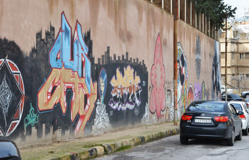 black car parked in front of a brown wall with street art on it, Amman