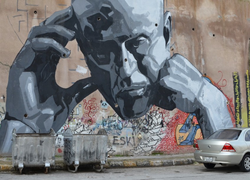 large mural in shades of grey, a man's head, chin on fist and trhe other hand resting on the other side of his head, on a wall, covering some older street art