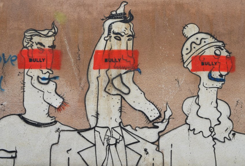 on a brown wall, three comical white characters, male, with exaggerated features drawn in black, each has an orange stripe through their face with the word Bully written on it