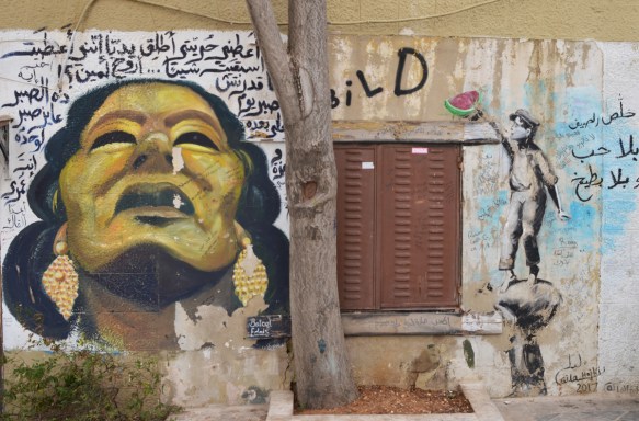 two street art paintings on the sides of a shuttered window. on the left, a gold coloured woman with braids looking to the sky, on the right, a person reaching for a piece of watermelon up on a shelf 