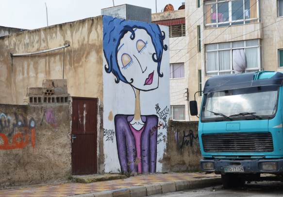 a turquoise truck is parked on a residential street in Amman beside a mural of woman with blue hair and a purple jacket, she has her eyes closed 
