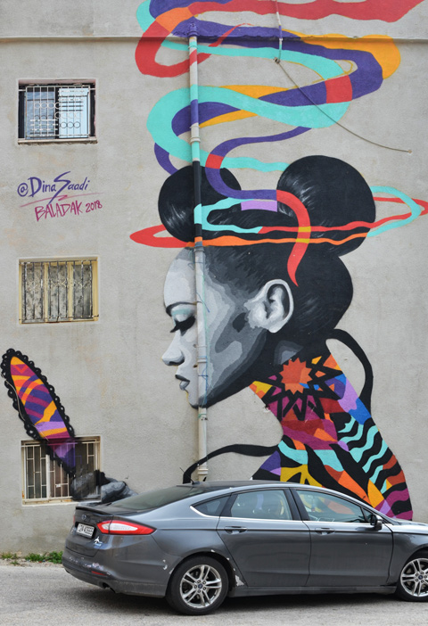 large mural in Amman of a woman looking in a hand held mirror, with hair tied in two buns and lots of colourful ribbons around her head 