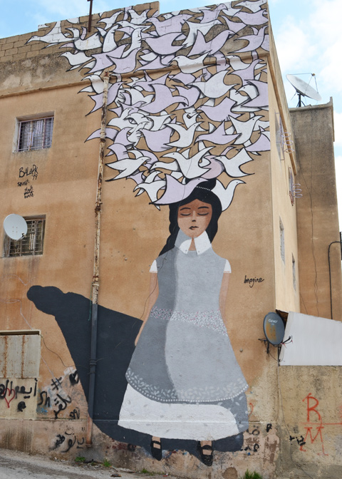 large mural of a woman wearing a grey top, eyes closed and dreaming of doves, the title is imagine and it was painted by sourati, on the side of a large building in Amman