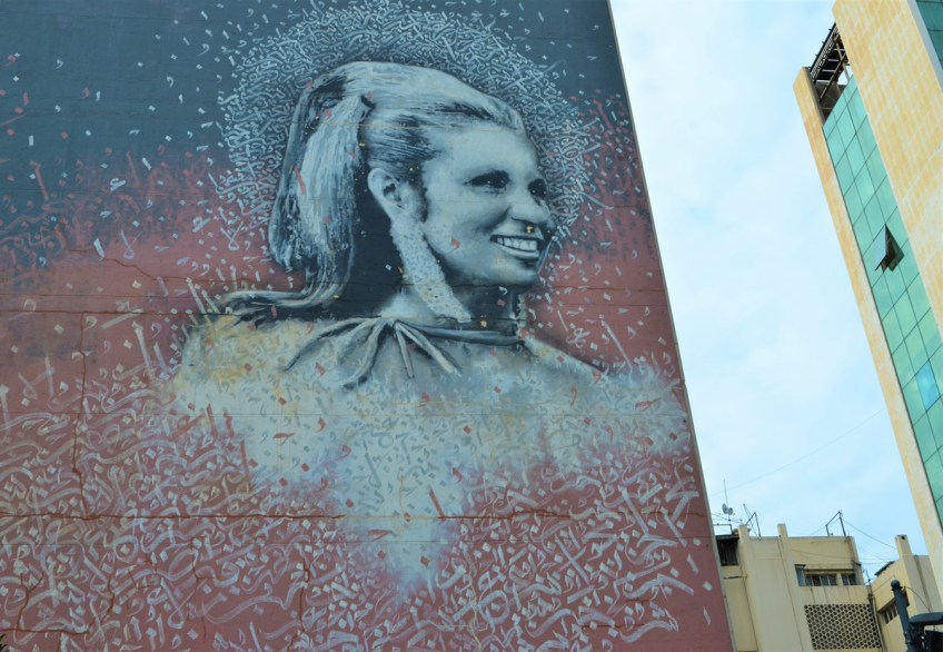 high on a wall, a large mural by Yazan Halwani of a woman's head and shoulders, surrounded by Arabic letters and words. she is seen in profile, smiling as she looks away