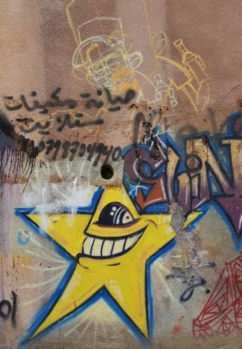street art on a wall in Amman, a yellow star with an open mouth with big teeth and one eye, looking to the left, Arabic writing and a phone number tag, remnants of a drawing street art 