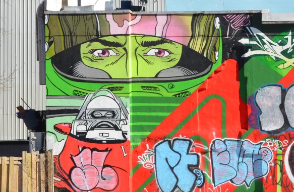 a mural that has been tagged over except for eyes on a green face behind a mask and a black and white figure in a cockpit