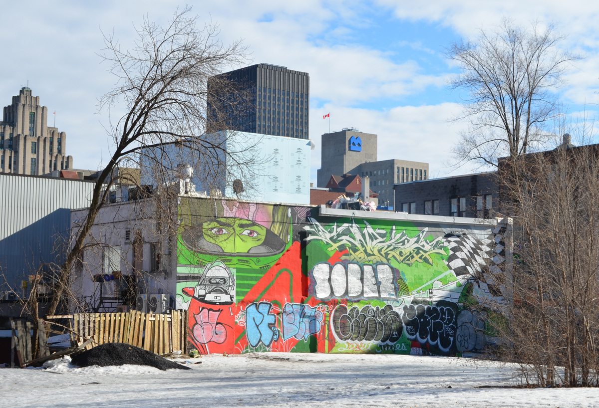 looking across snow covered park to a mural that has been tagged over except for eyes on a green face behind a mask and a black and white figure in a cockpit as well as black and white checkered flag in upper right corner, Montreal buildings behind the mural 
