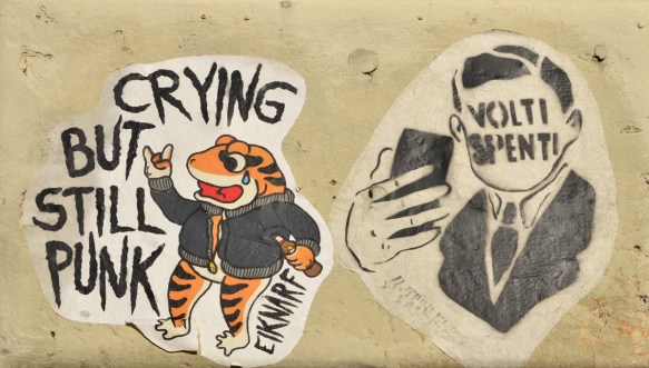 two slaps on a wall, one is a frog standing upright with a bottle in one hand and the words crying punk, by artist eiknarf. the other is a black stencil of a man with blank face taking a selfie on his phone. Instead of facial features he has the words volti spenti which are Italian 