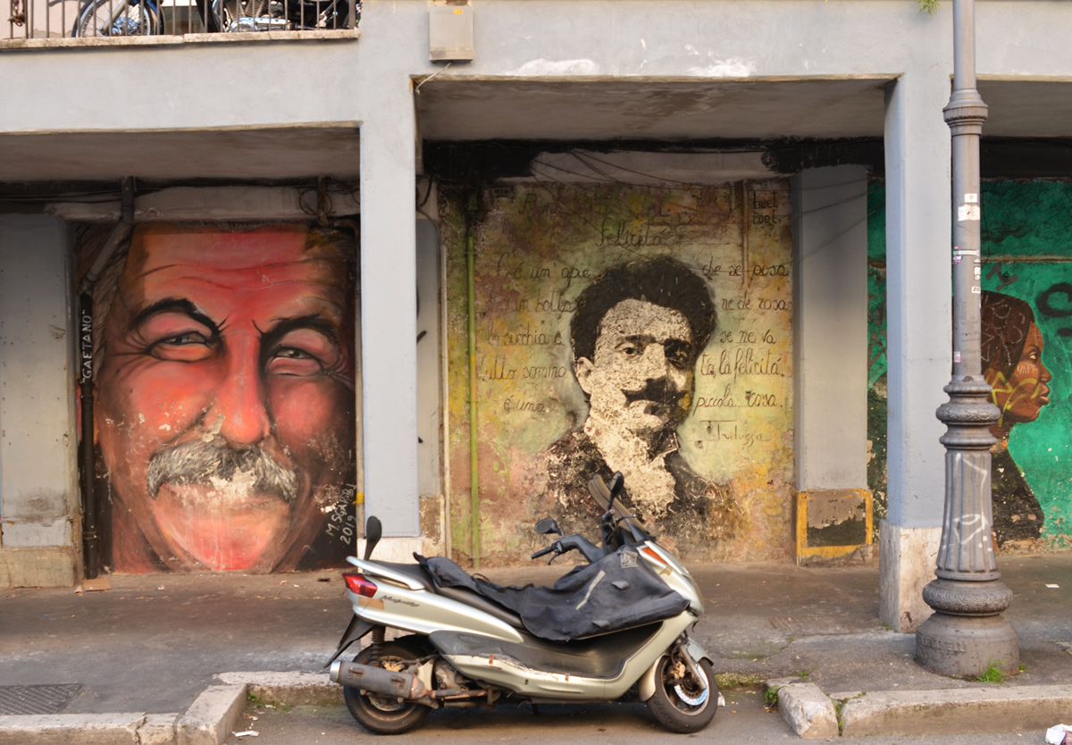 motorcycle parked in front of two mural of men's faces. one on the left is a just face, man with mustache called Gaetano. The other is an older man with Italian words written around him 