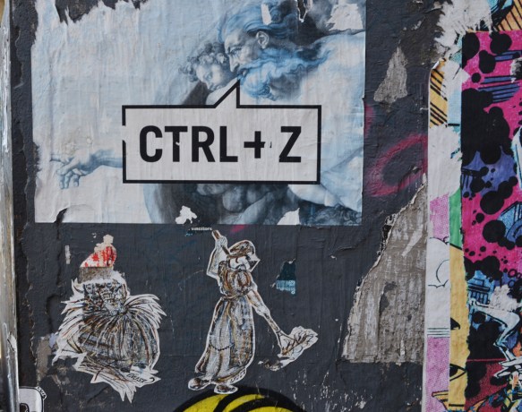 graffiti on a wall. one is a poster of Michelangelo's sistine chapel creation where god points finger at man, but this time, ctrl + z is written on top, the computer shortcut for undo. There are also two little drawings by L J S 57, women working, women in old fashioned clothing, one is sorting straw and the other is sweeping 