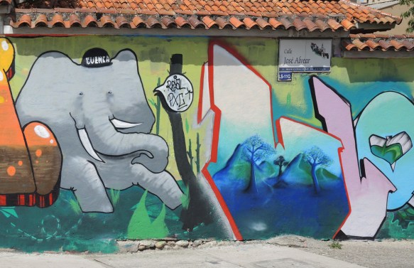 a grey elephant with a baseball cap that says Cuenca on it, also letters, possibly L and L, mural on a wall,