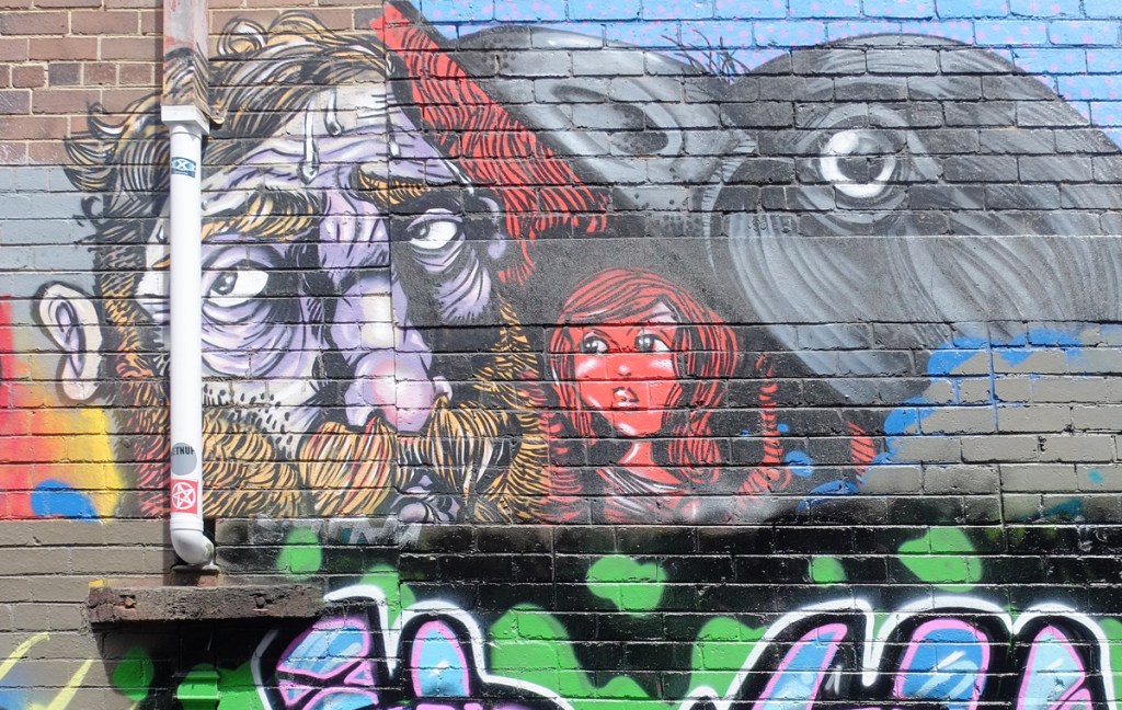 street art painting of a man's head, a woman's head and a bird's head, all above a text piece
