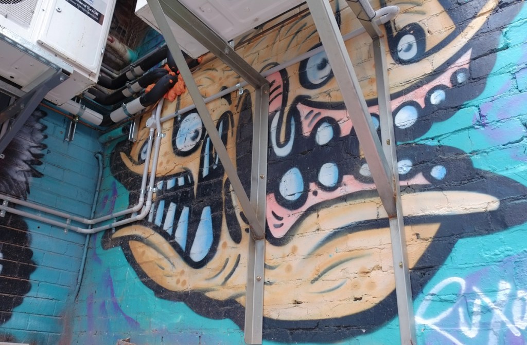street art in an alcove, up under air conditioning units and plumbing, looks like a sea creature with a big mouth and teeth