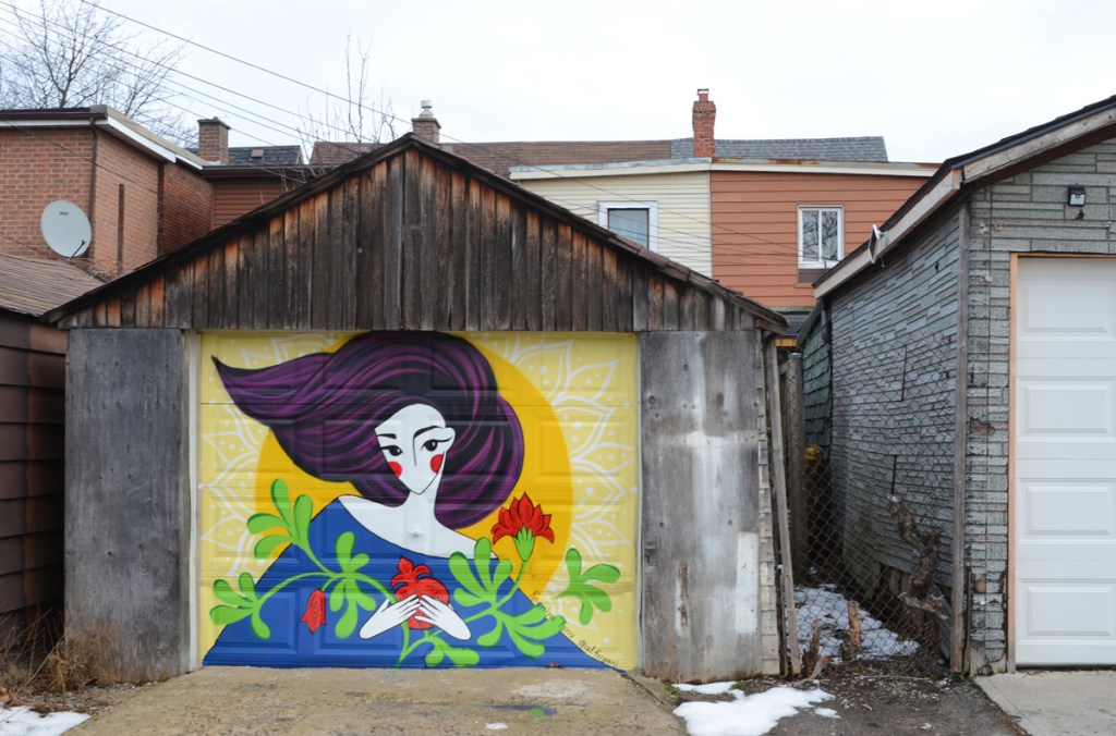 bright and colourful mural in an alley of a woman with long purplish black hair, very white skin, and a blue low necked top.  She's holding a pomegranate and two branches with green leaves and red flower