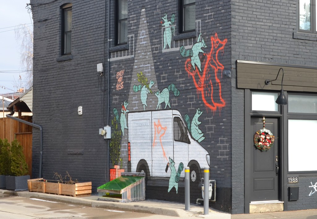 an emily may rose mural on the side of a dark grey building, showing raccoons playing around a white cube van, vandalism with orange spray paint