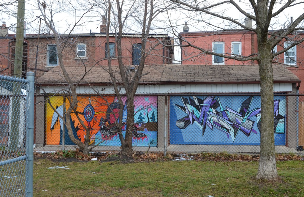 looking across a park in late December, two garages in an alley painted with murals, on the right is one in blues by mediah. On the left is mural mural by Que Rock, indigenous themes, loons on the water, mother earth