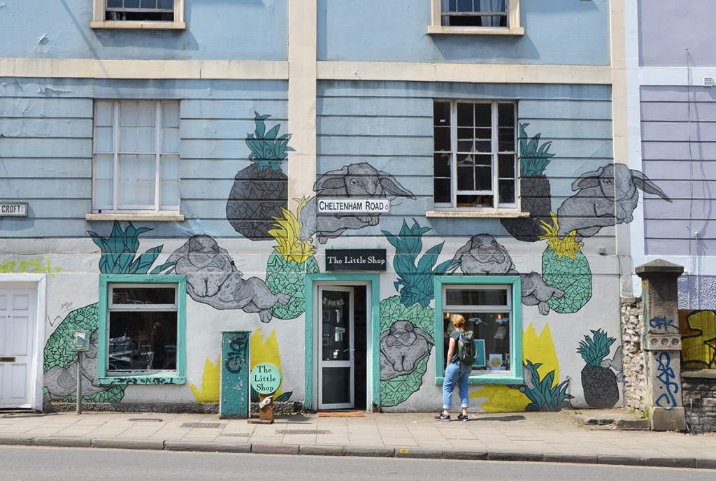 large mural by Lucas Antics on The Little Shop on Cheltenham Road in Bristol.  A woman is looking in the window of the store.  The mural is bunny rabbits and pineapples, 6 rabbits and 7 pineapples