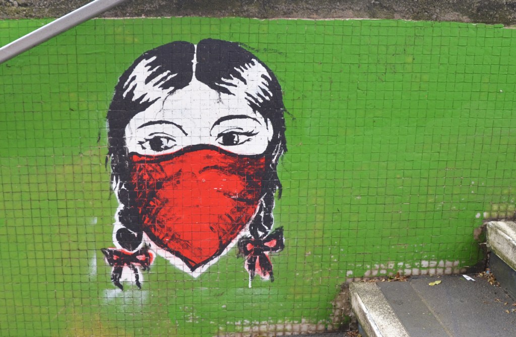graffiti of face of young girl with braids tied with ribbons, and wearing a red bandana mask over her nose and mouth, on a green tile wall beside stairs 