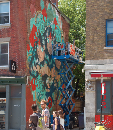 street artist nychos up on a lift and painting a large mural in Montreal as people walk by on the street