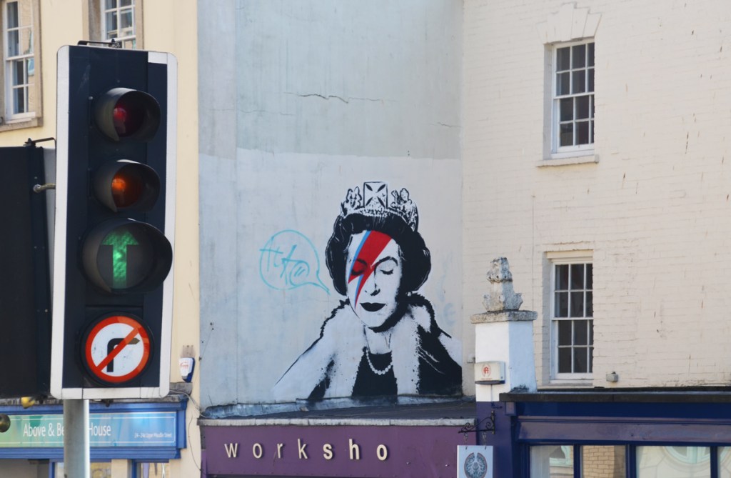 behind a traffic light, on the upper level of a building, on a pale grey wall, a Banksy street art.  A black and white Queen Elizabeth 2, younger woman, with a crown on her head.  She has a red and blue David Bowie lightning bolt across her face like the Aladdin Sane album cover of 1973 