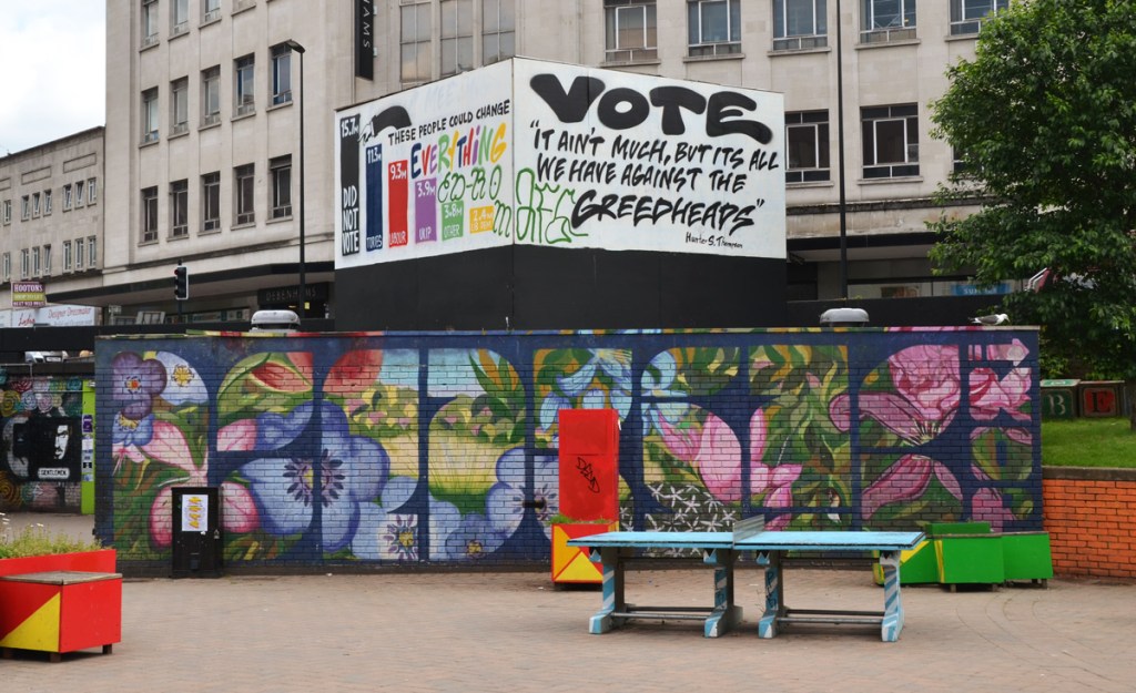 tables and planters at Bristol bearpit, park, with a mural of flowers.  In the background a sign, political poster, and in he background, Debenhams store