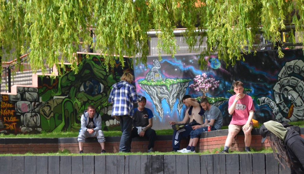 A group of young men sitting on benches in a park, with a street art wall behind them, space aliens and creatures