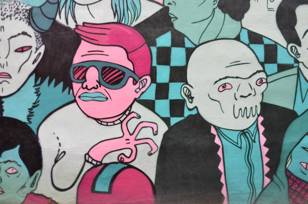 part of a mural  in a Montreal alley by Mono Sourcil, faces of people, including man with very pink face and dark pink hair, blue lips, another man with a face that looks more like a walrus 