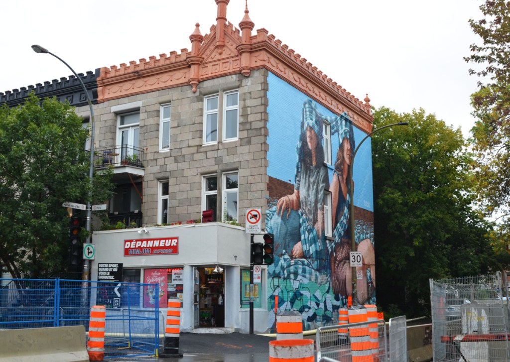 At the corner, beside a depanneur, a large mural of two seated women by Fintan Magee, one has blue and white checkered scarf over her face, the other has same scarf across the top of her head. 