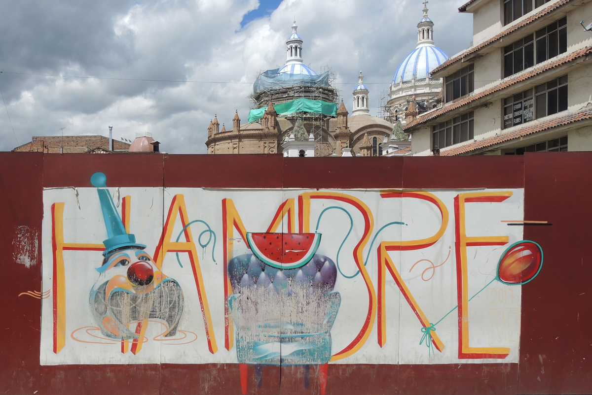 mural on hoardings with historic church in the background, word hambre with 4 objects, a clown head, armchair, watermelon piece, and a red balloon on a string