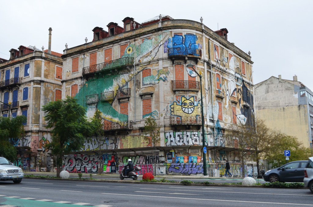 large abandoned old building on Av Fontes Pereira Melo in Lisboa that is covered with street art including a large green crocodile or alligator, a yellow cat on a balcony and large birds that cover one side of the building