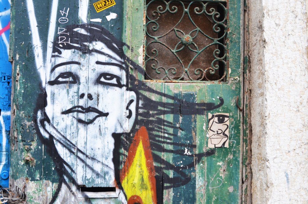 street art painting of a young woman on a door, she's looking upward, beside a small square window with a metal grille