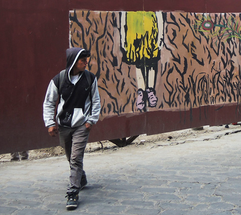 a young man in a hoodie is walking past murals on hoardings, picture on mural is of a person wearing purple shoes, Cuenca Ecuador