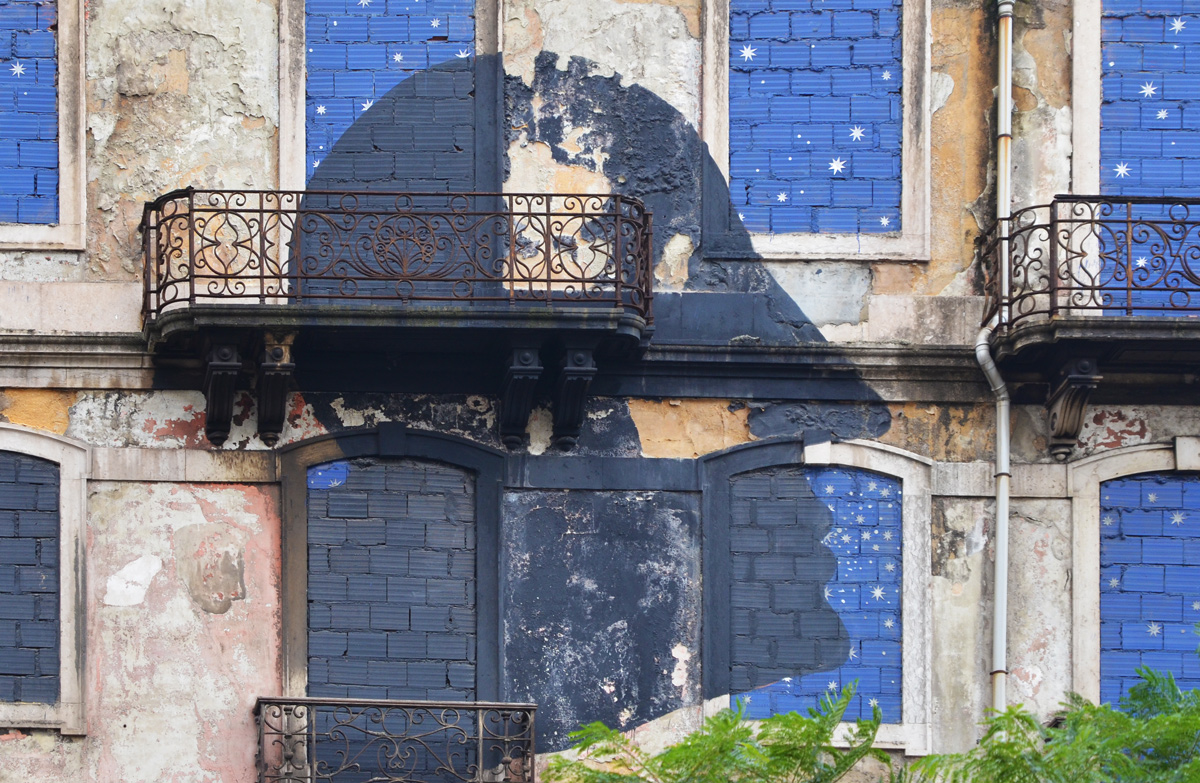 part of a mural on a Lisbon building, black silhouette of a man's head, windows painted night sky blue, balconies with wrought iron railings