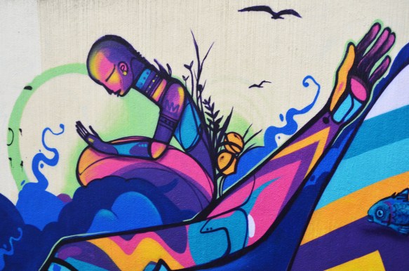 mural street art in bright jewel tones, purple, dark pink, turquoise of a person sitting looking at their hand, also a long arm with fingers