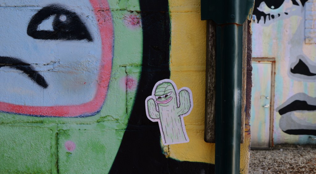 a small sticker of a cactus with a face, stuck on a mural beside a face