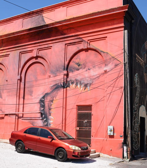 a car parked in front of a mural that is the same orangish red that the background of the mural is, a large shark with mouth open and many teeth 