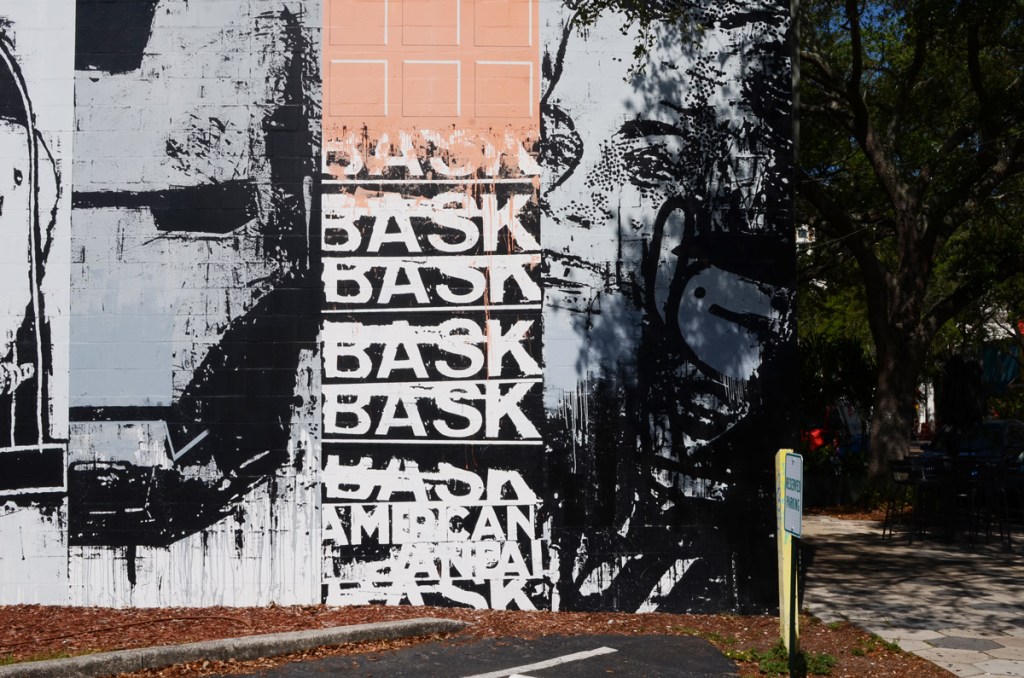 part of a mural by bask, in shades of grey, collage of different pictures, the word bask in capital letters written many times, an iron man mask, a woman's face